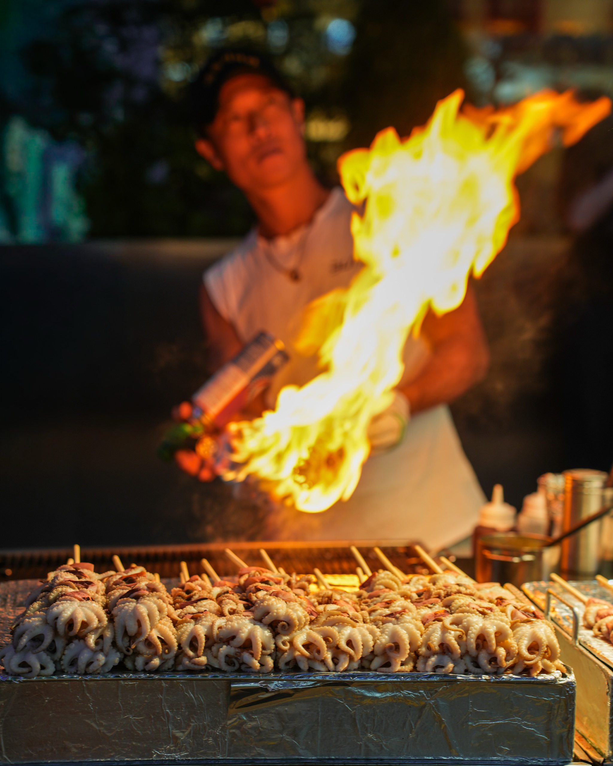 A street food vendor torching skewers over an open flame at night